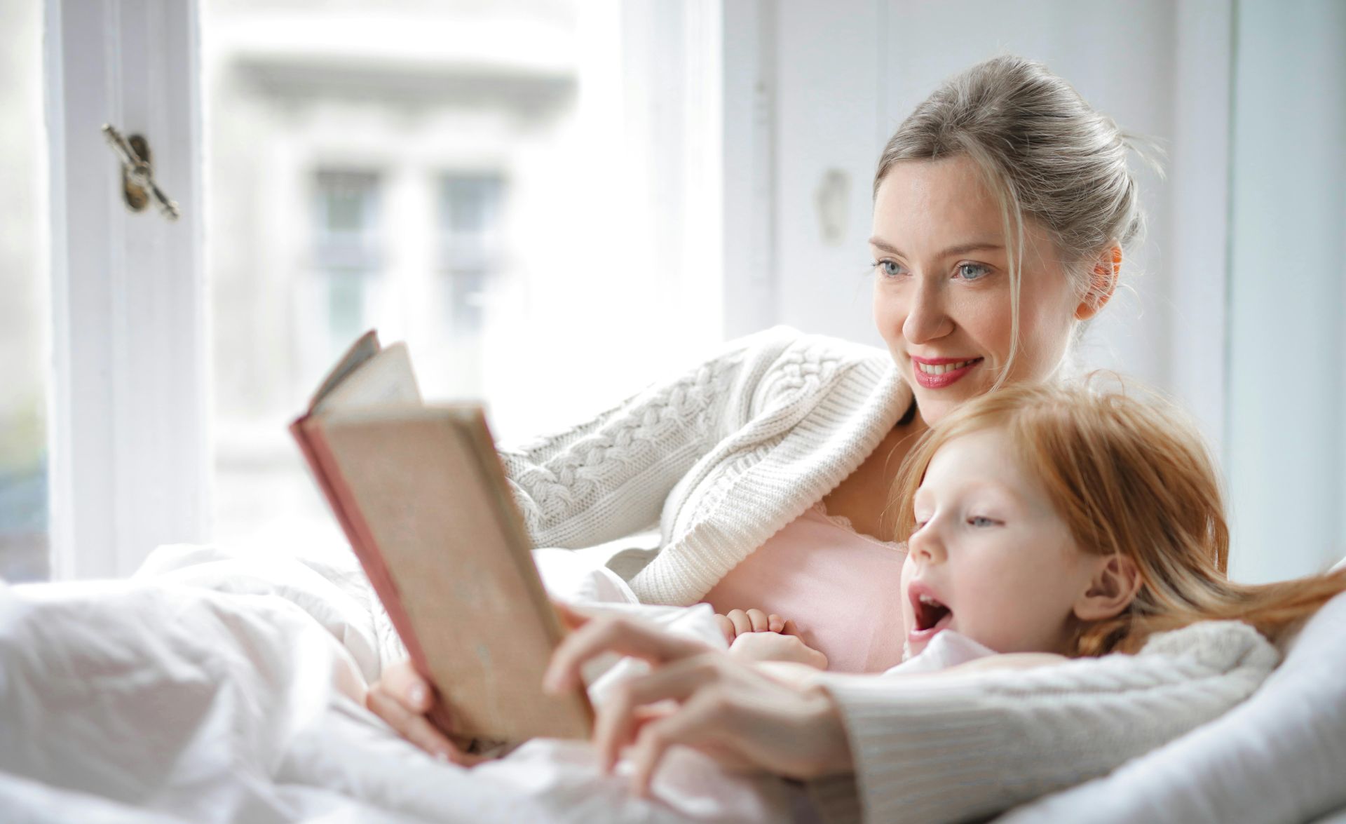 Cheerful young woman hugging cute little girl and reading book together while lying in soft bed in light bedroom at home in daytime